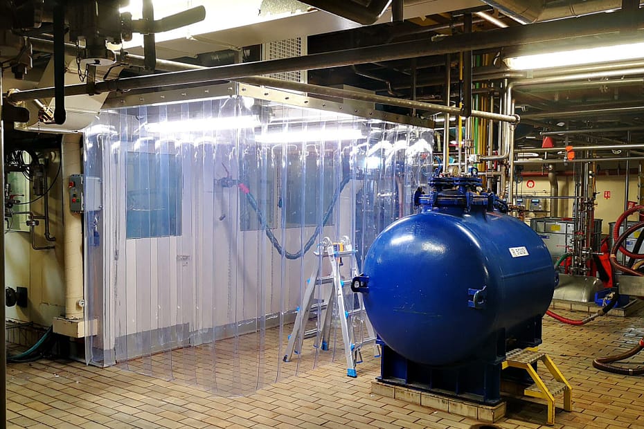 Industrial equipment room with a large blue spherical tank, transparent plastic curtain, and an overhead network of pipes and valves.